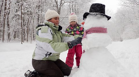 A mother and daughter having fun building a snowman. The woodland area they are in is covered in a blanket of snow and surrounded by trees. The snowman wears a black hat, red and white striped scarf and has a carrot for a nose. The mum has a green waterproof coat and white hat, while her daughter has a white hat with a pink bobble, a multicoloured grey, green and pink coat and dark pink pants on