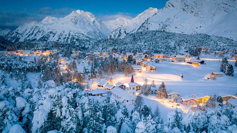 A twilight, an alpine village is surrounded by snowy woodland and mountains. A the centre of the village is houses, a church and other buildings and all their rooftops are covered in snow. Light is beaming around them, coming from the windows,as the night gets darker
