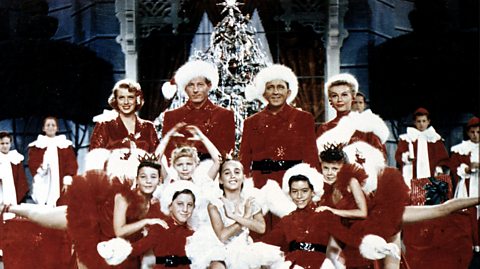 A scene from 1954 Christmas musical film White Christmas. Pictured, Rosemary Clooney, Danny Kaye, Bing Crosby and Vera-Ellen in front of a Christmas tree, all wearing red outfits with white fur trim. They are surrounded by children in similar outfits who gather and pose in front of them, at the centre, one little girl is dressed as an angel