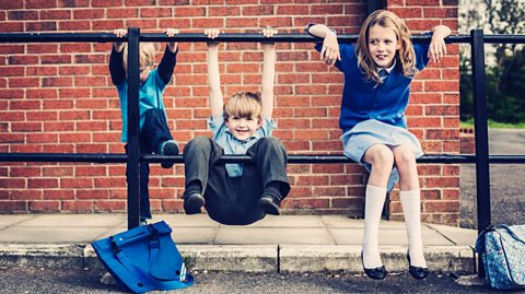 three children in blue school uniforms are sitting on and hanging off a railing beside a brick building - probably waiting for their parents at school.