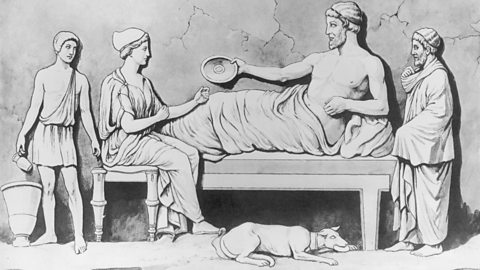 Four people are gathered around a couch. A man is lying down and holding a round plate, while a woman sits on a stool nearby. Two others stand close, one holding a jug. A dog is resting on the floor in front of them.