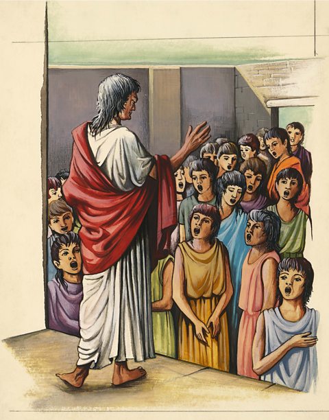 A man wearing a white robe with a red cloth draped over one shoulder is standing and speaking to a group of children. The children are listening closely, and they are wearing colorful clothes like yellow, blue, green, and purple.