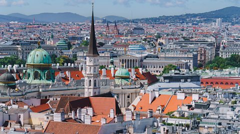 An aerial view of Vienna, the capital of Austria, in the sunshine. Images shows lots of city centre buildings with traditional orange Terracotta roofs, a tall spire of the cathedral and some domed buildings
