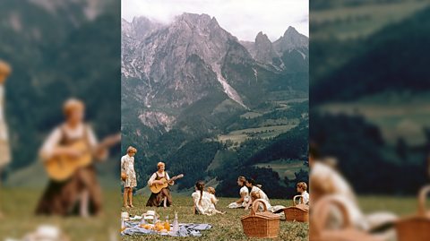 Julie Andrews and the cast of The Sound of Music filming in Austria. She sits in a field playing the guitar and wearing a brown pinafore style dress with a long sleeved white top underneath. The backdrop is the Alps mountains and the children sit around her all having a picnic