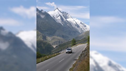 The highest mountain of Austria, the Großglockner. Image shows the mountain, covered in a dusting of snow, in the background and a road surrounded by green land as a grey car drives down it