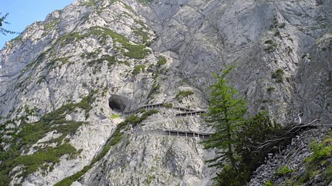 The entrance to Eisriesenwelt - the world's largest ice cave, in Austria. The image shows a hole inside a large, rocky mountain, which is covered in some greenery and tracks leading up it for tourists to enter