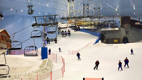 Inside a massive indoor ski experience, with slopes of snow and skiers descending down them. To the left is a large chairlift taking groups of two and three people to the top