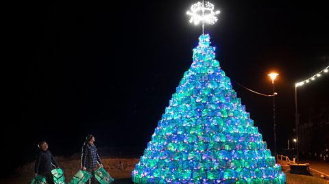 Christmas tree made from stacked illuminated fishing creels glowing in blue and green, topped with a bright star, displayed outdoors at night with two people holding matching lighted decorations.