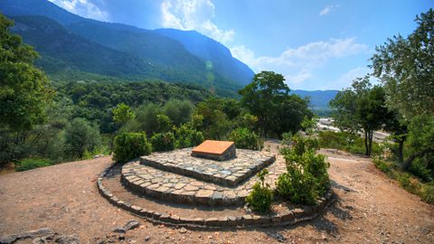 A small stone platform in the middle, surrounded by green bushes and trees. Behind it are big mountains covered in forests, and the sky is bright blue with white clouds.