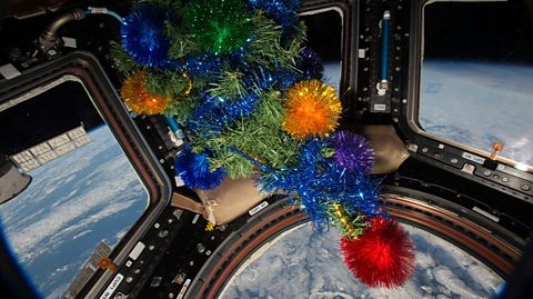 A mini Christmas tree with bright shiny tinsel, in the International Space Station's Cupola module as it orbits Earth. Through the windows can see Earth down below 