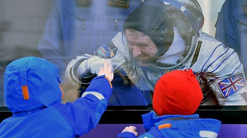 Astronaut Tim Peake, in his space suit, fist-bumps one of his two young sons through the bus window. We see the back of his two boys in bright blue snow suits, one with a red woolly hat