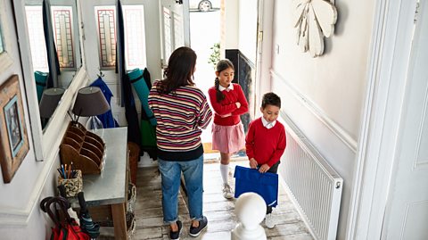 Mother opens door to 2 children in red school uniforms as they enter hallway of their home on return from school.