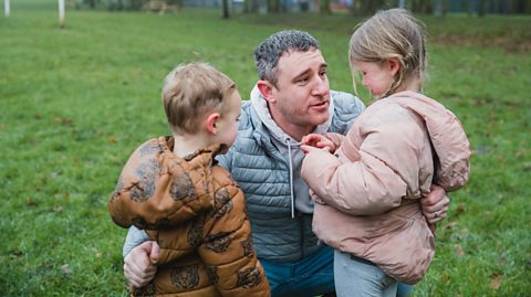 Dad comforts his primary school age son and daughter in the park as the daughter is upset.