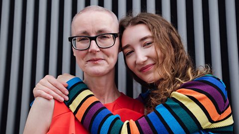 Mum undergoing cancer treatment is hugged by her late teen daughter. Both smile for the camera.