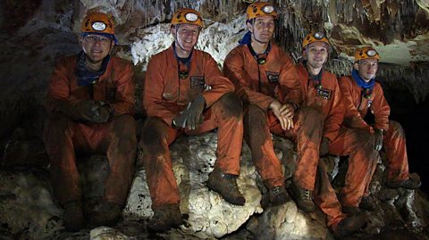 Five men wearing orange jumpsuits, hard hats and headtorches sit in a cave. Their boots, clothing and faces are muddy.