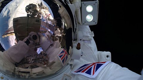 Tim Peake wearing full space suit and helmet as he completed a spacewalk, take a selfie in the reflection so we see the ISS and Earth behind him