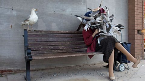 Art installation at Banksy’s Dismaland showing a mannequin seated on a bench being swarmed by seagulls, with another seagull perched on the bench and a shopping trolley nearby