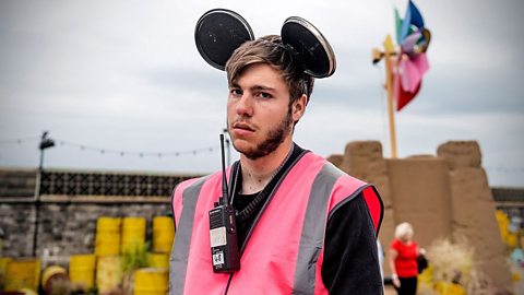 Event staff member at Banksy’s Dismaland wearing a bright pink high-visibility vest and black Mickey Mouse-style ears, with a walkie-talkie clipped to the vest, standing in front of a sandcastle installation and colourful flags.
