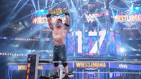 WWE wrestler John Cena holds the WWE Championship above his hide while climbing the ropes in the corner of a wrestling ring. Wrestlemania signs are in the background as is the number 17
