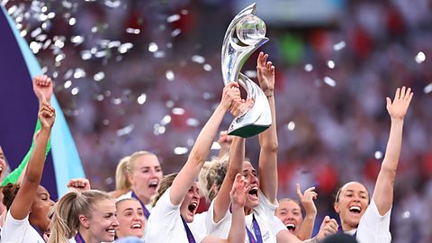 Ellen White and Jill Scott jointly lift the trophy in the air after winning Euro 2022 for England. Their teammates are around them, arms aloft, with ticker tape falling in the background