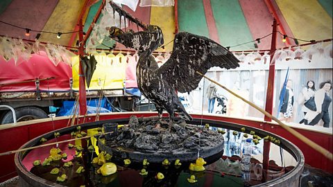 Art installation at Banksy’s Dismaland featuring a blackened pelican sculpture standing on debris in a circular water tank, surrounded by floating yellow rubber ducks under a colourful striped canopy, with people walking past it behind.