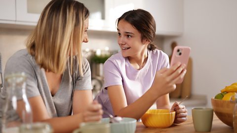 Mother and daughter are sitting at the kitchen table, having breakfast and looking at social media on a smartphone