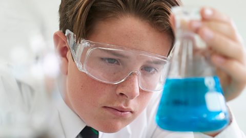 School science student wearing goggles and holding up a flask with blue liquid