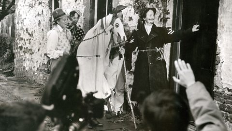 A black and white photograph of the Mari Lwyd tradition in Wales. The Mari Lwyd figure is being led to a door - in the traditional set up of a horse skull, white sheet and being led by others