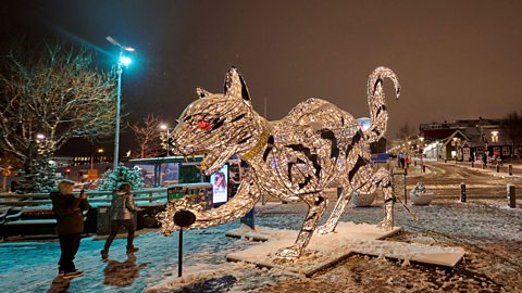 A person takes a photograph of a large Yule Cat festive statue that is lit up. It's eyes are red and it's in a position ready to pounce