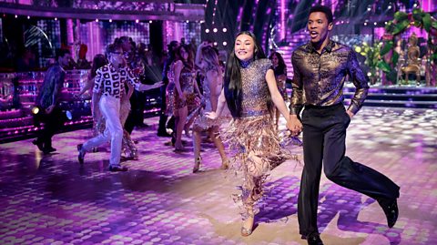 Couples performing a lively ballroom dance routine on a brightly lit stage with purple and pink spotlights, wearing glittering costumes and metallic shirts, surrounded by other dancers in a glamorous setting.