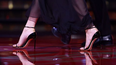 Close-up of elegant black stiletto heels on a glossy red dance floor, with sheer flowing fabric and a partner’s shoes visible in the background during a ballroom performance