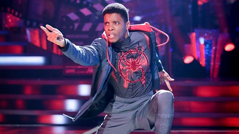 Rhys Stephenson kneeling on a stage in a dynamic dance pose, wearing a Spider-Man themed shirt, dark pants, a long coat, and red sneakers, with vibrant red and purple stage lighting and bold signage in the background.