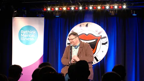 A man performs at Men's Minds Matter suicide prevention comedy course – a sign next to him that reads "Rethink Mental Illness" (Credit: Joe Bor)