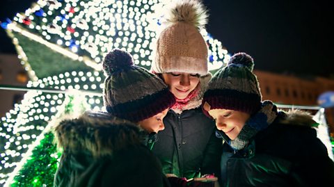 Two kids and their mother wearing hats checking the smartphone with Christmas lights and decorations visible in the background