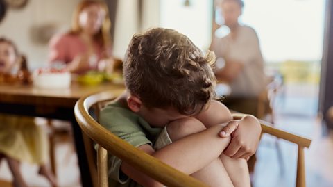 child huddled in chair upset with family blurred in background at dinner table