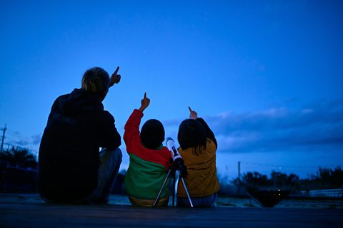 Silhouette of father and two children sitting on the ground and pointing to the sky in search of stars