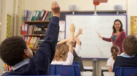A female teacher stands at the front of a classroom smiling, as her pen points to the whiteboard. She is wearing a blue top with a pink cardigan over it and has brunette hair. Next to the whiteboard is a large shelf with colourful folders and books on it. In the forefront of the images is the back of children's heads as they face their teacher and raise their hands to get her attention. The classroom children wear navy blue jumpers and blue polos, as well as white tops and grey pinafores