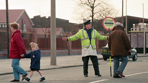 A little girl with blonde hair wearing a navy blue puffy coat crosses the road holding hands with a woman with short curly brown hair, who wears light blue jeans and a red waterproof coat. They are assisted by a lollipop man, holding a vibrant lollipop stick and fluorescent yellow high-vis jacket, black trousers and a black cap. Also crossing the road is an older man with grey hair wearing a big brown coat on and jeans. On the left hand side of the road, we see red brick houses, a green car driving and a yellow bus shelter in the distance