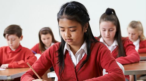 Five children, three girls and a boy, sit on desks lined up under exam conditions. They all wear red cardigans and jumpers for uniform and are writing in exam papers using wooden pencils