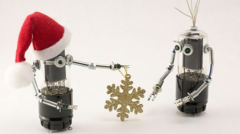 Two simple robots meet, one wearing a red Santa hat gives the other a gold snowflake decoration