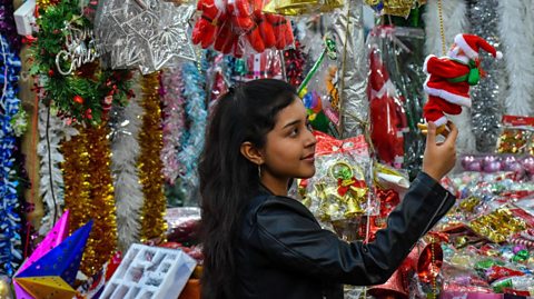 A young woman holds aloft a small Father Christmas toy at a market stall covered in tinsel and sparkly decorations