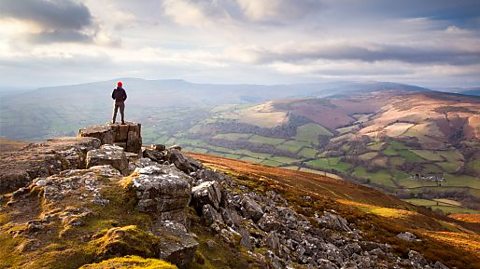 A small and distant figure can be seen stood against a mountain sunset in the Brecon Beacons National Park, Wales. The landscape looks cloudy and vast