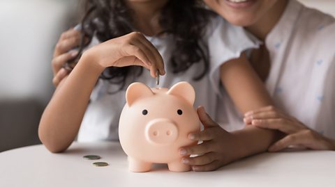 Little girl sitting next to her mother and putting a coin into a piggy bank