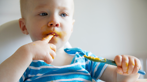 A baby messily eats mashed food from a plastic spoon.