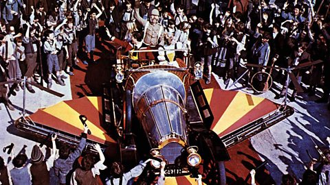 Dick van Dyke as Caractacus stood in the back of Chitty Chitty Bang Bang, surrounded by cheering people