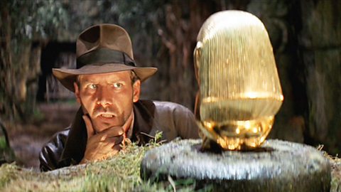A still from Raiders of the Lost Ark. In trademark hat, Indiana Jones strokes his chin while contemplating taking the golden idol