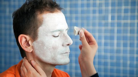 A clown as he starts to get his face painted. The man has brown hair and wears an orange top. His face is painted white as a hand dabs more on with a sponge