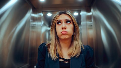 A blonde woman wearing a black top and blue blazer holds her breath and looks uncomfortable in an elevator as she does not like confined spaces