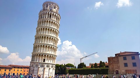 Tourists gather around the bottom of the Leaning Tower of Pisa and take photos of the medieval structure, which has a white and grey marble facade and a striking tilt