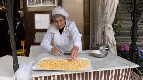 A woman wearing a white chef jacket and hat making pasta in Italy. In front of her is a tray of pasta and a bowl of ingredients